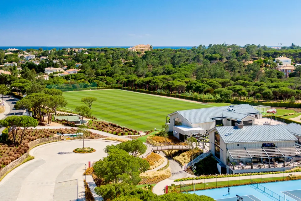 Aerial view of The Campus, Quinta do Lago — the training pitch, clubhouse and tennis courts among pine woods, with the sea beyond